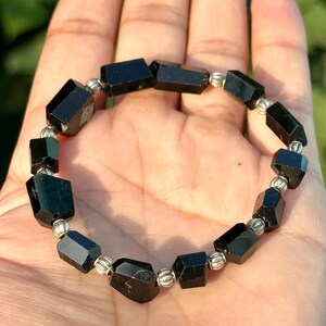 以下が含まれることがあります： 黒トルマリンのブレスレット。長方形のファセットビーズと銀色の金属ビーズが組み合わされています。ブレスレットは手の上にあり、背景はぼやけた緑色です。