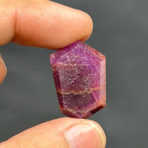 May include: A faceted, hexagonal ruby gemstone with a deep magenta hue. The gem is held between two fingers, showcasing its polished surfaces and natural inclusions. The stone's color varies from deep red to a lighter pink.