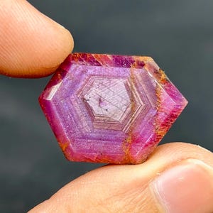Könnte beinhalten: Ein facettierter, sechseckiger Rubin-Edelstein. Der Edelstein zeigt eine tiefrosa und violette Farbe mit inneren geometrischen Mustern. Die Kanten weisen Farbtöne von Orange und Braun auf.