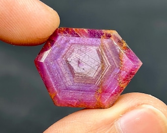 May include: A faceted, hexagonal ruby gemstone. The gem displays a deep pink and purple colour with internal geometric patterns. The edges show hints of orange and brown.
