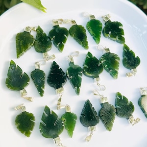 Puede incluir: Una colección de colgantes de hojas de jade verde con herrajes de metal plateado. Los colgantes están tallados en varias formas y tamaños de hojas, exhibidos sobre una superficie blanca. El jade tiene una calidad translúcida.