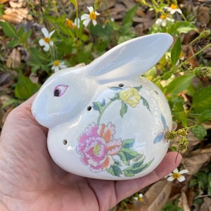 Könnte beinhalten: Eine weiße Keramik-Hasenfigur mit Blumendesigns in Rosa, Gelb und Grün. Der Hase hat eine rosa Nase und schwarze Augen.