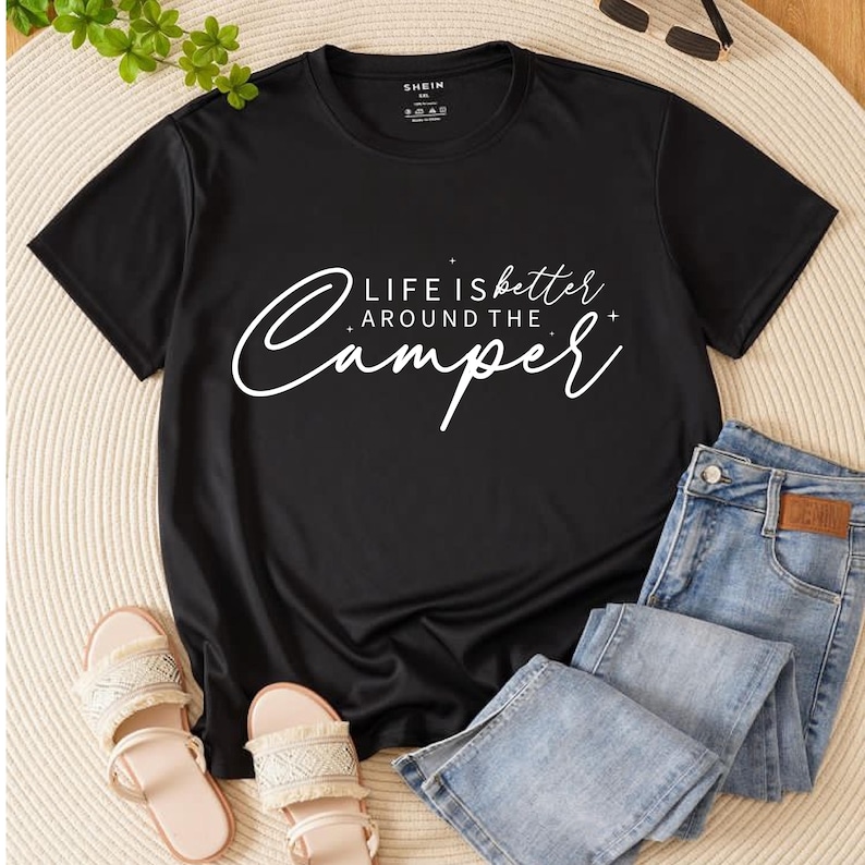 May include: Black t-shirt with the text "LIFE is better AROUND THE Camper" in white script. The shirt is paired with light wash denim jeans and beige sandals. Sunglasses are visible in the background.