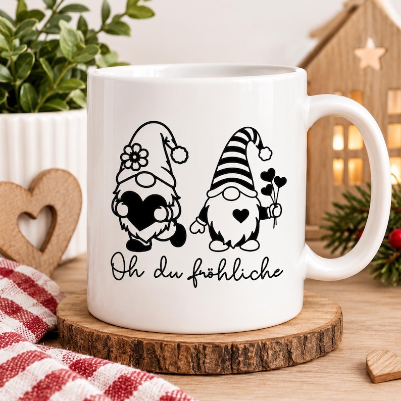 Puede incluir: Una taza de cer&aacute;mica blanca con un gr&aacute;fico negro de dos gnomos, uno con un sombrero adornado con una flor y sosteniendo un coraz&oacute;n, el otro con un sombrero a rayas sosteniendo tres corazones peque&ntilde;os, con el texto Oh du fr&ouml;hliche en negro.