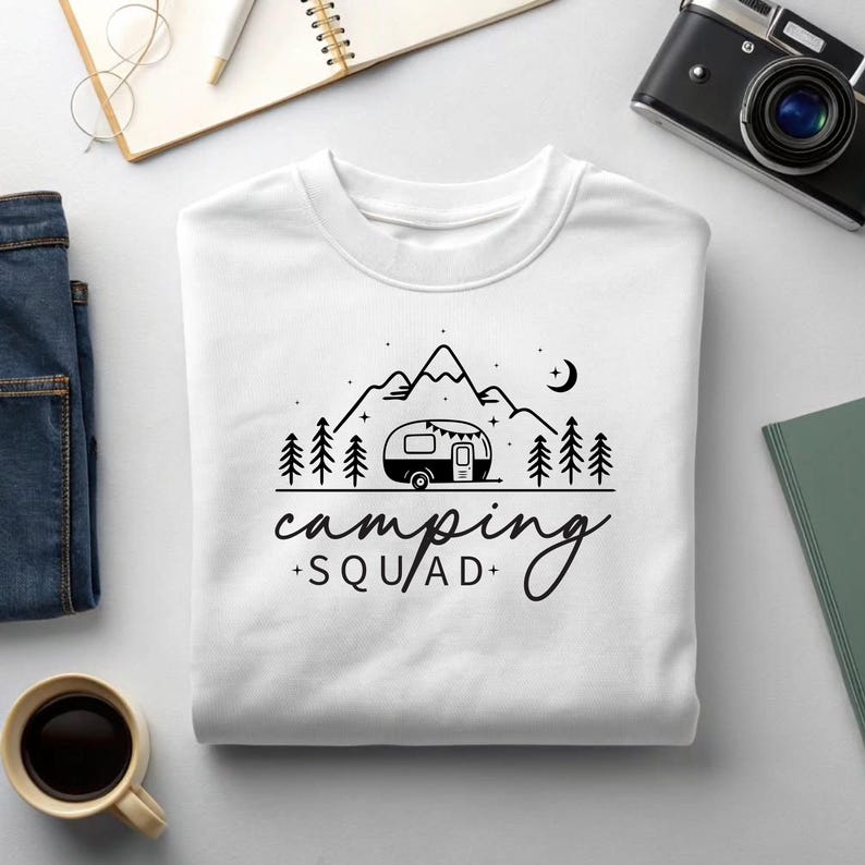 May include: White sweatshirt with a black graphic of a camper, mountains, trees, and a crescent moon. The text "camping + SQUAD+" is below the graphic. The sweatshirt is folded and surrounded by a notebook, camera, and coffee cup.