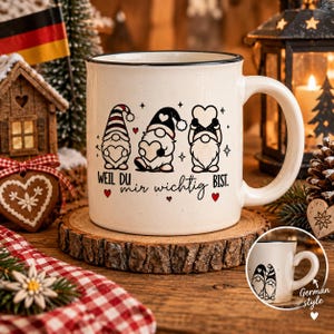 Puede incluir: Una taza de cer&aacute;mica color crema con borde negro, que presenta un gr&aacute;fico de tres gnomos sosteniendo corazones y el texto en alem&aacute;n "WEIL DU mir wichtig BIST." La taza se muestra sobre una rodaja de madera, con una peque&ntilde;a imagen insertada.