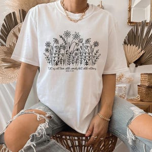 May include: White t-shirt featuring a black floral design with the text "Let us not love with words but with actions John 3:18". The shirt has short sleeves and a relaxed fit. The design includes various flowers and foliage.
