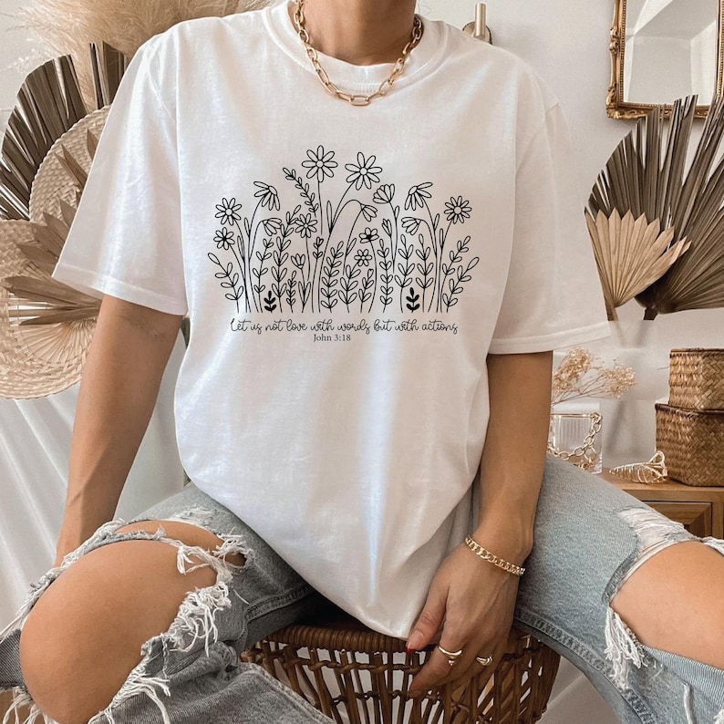May include: White t-shirt featuring a black floral design with the text "Let us not love with words but with actions John 3:18". The shirt has short sleeves and a relaxed fit. The design includes various flowers and foliage.