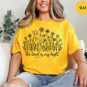 May include: Mustard yellow t-shirt with a black floral design and the text "The Lord is my light" from Psalm 27:1. The shirt has short sleeves and a relaxed fit. A gold circle is in the upper right corner.