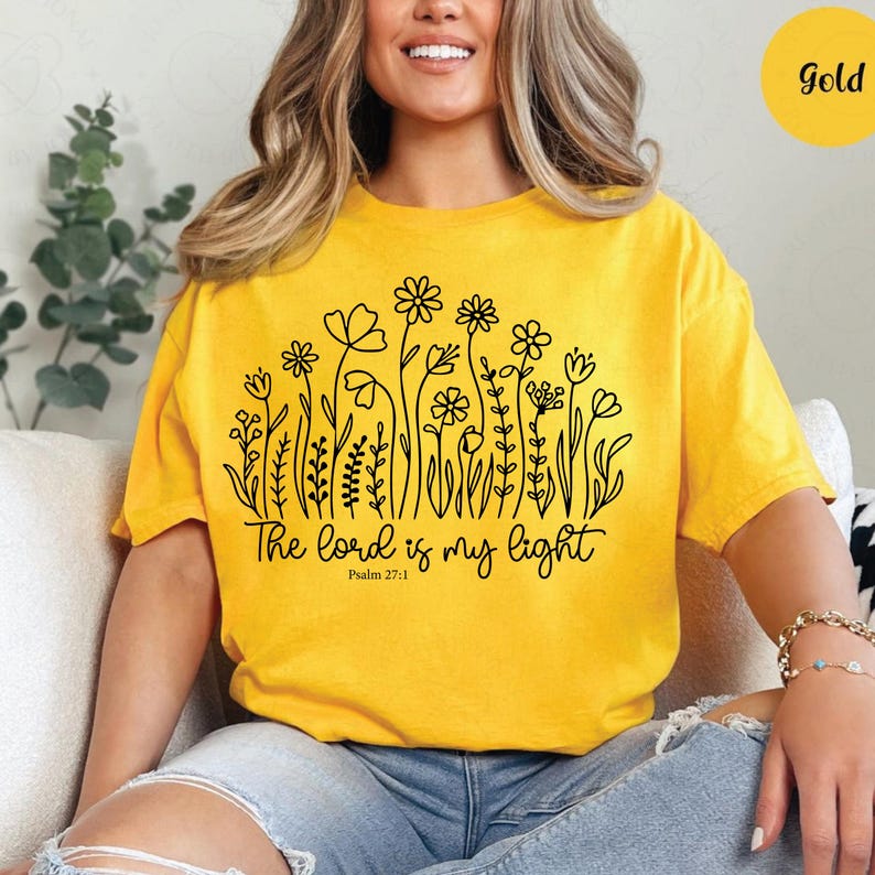 May include: Mustard yellow t-shirt with a black floral design and the text "The Lord is my light" from Psalm 27:1. The shirt has short sleeves and a relaxed fit. A gold circle is in the upper right corner.