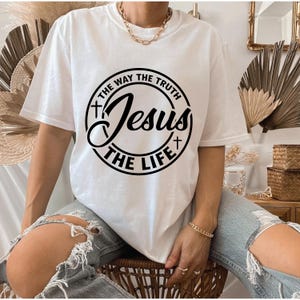Puede incluir: Camiseta blanca con un gr&aacute;fico circular negro que dice "Jesus The Way The Truth The Life". La camiseta tiene mangas cortas y cuello redondo. La persona lleva vaqueros azules rotos y joyas doradas.