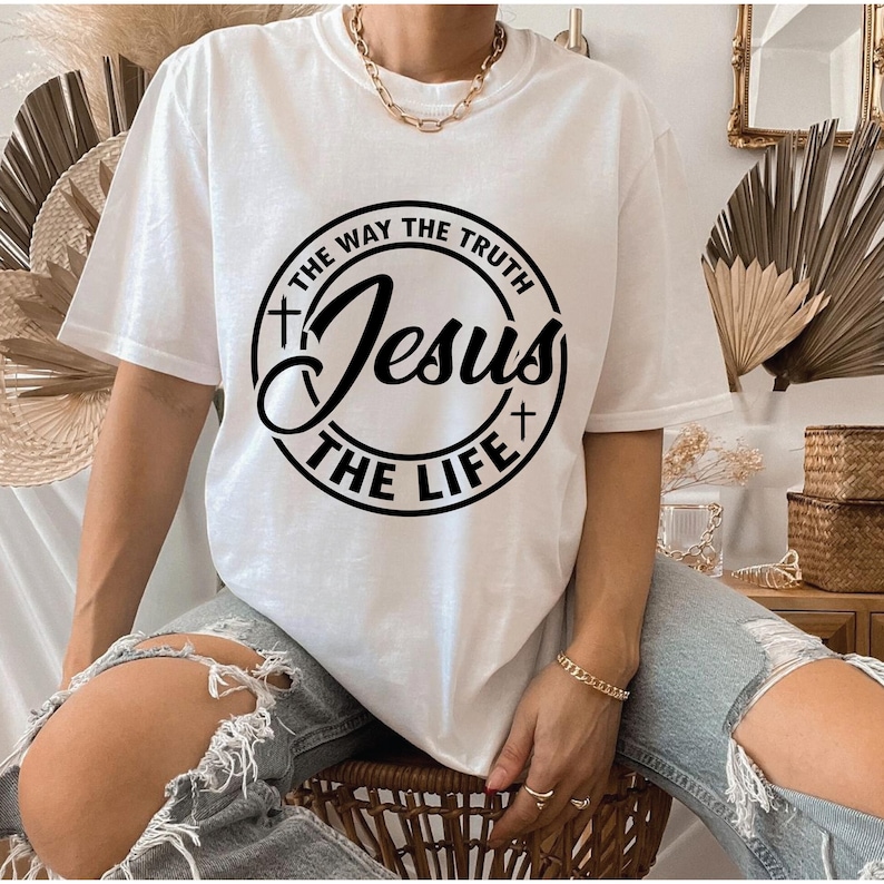 Puede incluir: Camiseta blanca con un gr&aacute;fico circular negro que dice "Jesus The Way The Truth The Life". La camiseta tiene mangas cortas y cuello redondo. La persona lleva vaqueros azules rotos y joyas doradas.