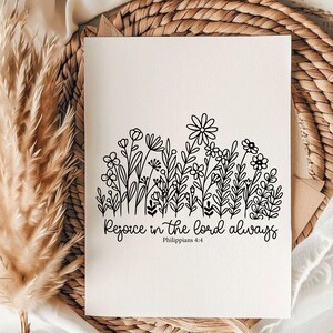 May include: A cream-colored card features a black floral illustration at the top and the text "Rejoice in the Lord always" with "Philippians 4:4" below. The card is set against a woven background with dried plants.