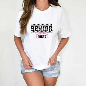 May include: White t-shirt with the word "SENIOR" in black block letters over a black and white checkered pattern. Below the word "SENIOR" is a pink bow and the number "2027". The shirt is a casual style.