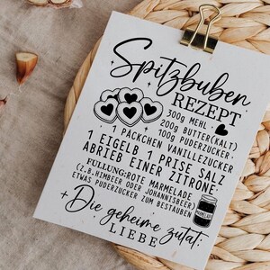 May include: A white recipe card with the title "Spitzbuben Rezept" in elegant script, held by a gold clip. The card features a list of ingredients and instructions for making Spitzbuben cookies, including measurements in grams.