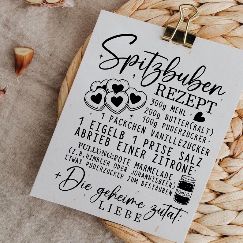May include: A white recipe card with the title "Spitzbuben Rezept" in elegant script, held by a gold clip. The card features a list of ingredients and instructions for making Spitzbuben cookies, including measurements in grams.