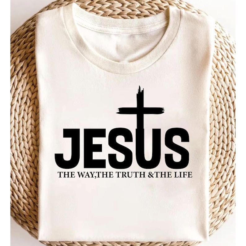 Puede incluir: Camiseta color crema con la palabra "JESUS" en letras negras en negrita y una cruz negra. Debajo, el texto "THE WAY, THE TRUTH & THE LIFE" en una fuente m&aacute;s peque&ntilde;a. La camiseta se muestra sobre una superficie tejida.