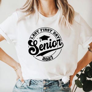 May include: White t-shirt with a black circular graphic that reads "Last First Day Senior 2021". The word "Senior" is in a bold, cursive font. A graduation cap is above the word "Senior".