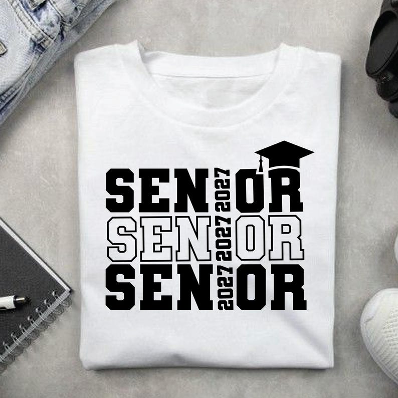 May include: White t-shirt with the word "SENIOR" stacked three times in bold black letters. The year "2027" is printed vertically on the right side of the word "SENIOR". A black graduation cap is above the last "OR".