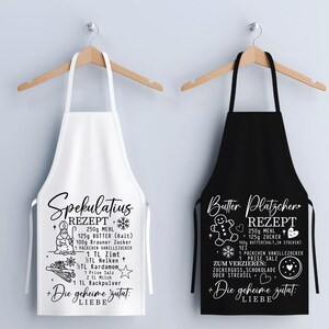 May include: Two aprons hanging on wooden hangers. One is white with a black recipe for "Spekulatius" cookies, and the other is black with a white recipe for "Butter Pl&auml;tzchen" cookies. Both aprons have a recipe list and the phrase "Die geheime Zutat: LIEBE".
