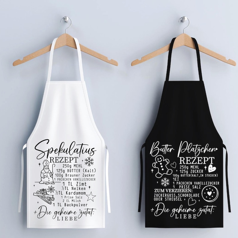 May include: Two aprons hanging on wooden hangers. One is white with a black recipe for "Spekulatius" cookies, and the other is black with a white recipe for "Butter Pl&auml;tzchen" cookies. Both aprons have a recipe list and the phrase "Die geheime Zutat: LIEBE".