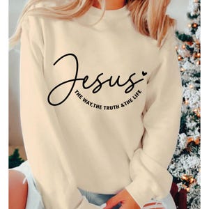 Puede incluir: Sudadera color crema con la palabra "Jesus" en cursiva, acompa&ntilde;ada de la frase "The Way, The Truth & The Life" en letra m&aacute;s peque&ntilde;a. Un peque&ntilde;o coraz&oacute;n est&aacute; junto a la palabra "Jesus".
