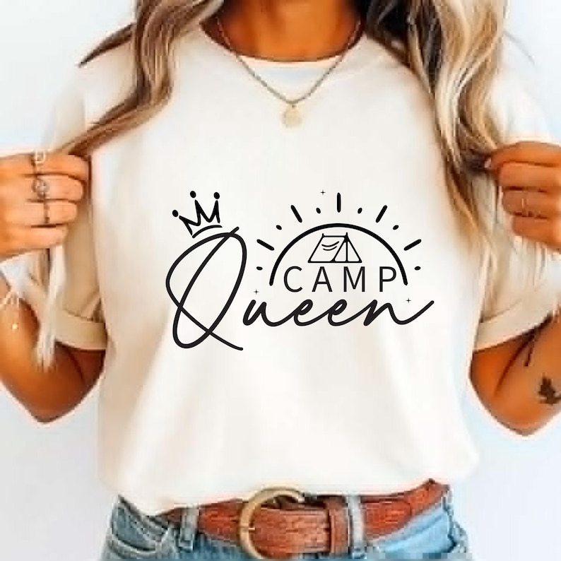 May include: Cream-colored t-shirt featuring a black graphic design. The design includes the word "Queen" in a cursive font, a crown, and the word "Camp" with a tent illustration. The shirt is a casual wear item.
