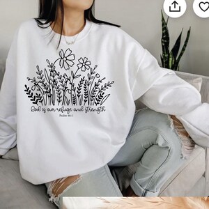 May include: White sweatshirt featuring a black floral design with the text "God is our refuge and strength" and "Psalm 46:1". The sweatshirt has a crew neck and long sleeves. The design includes flowers, leaves, and stems.