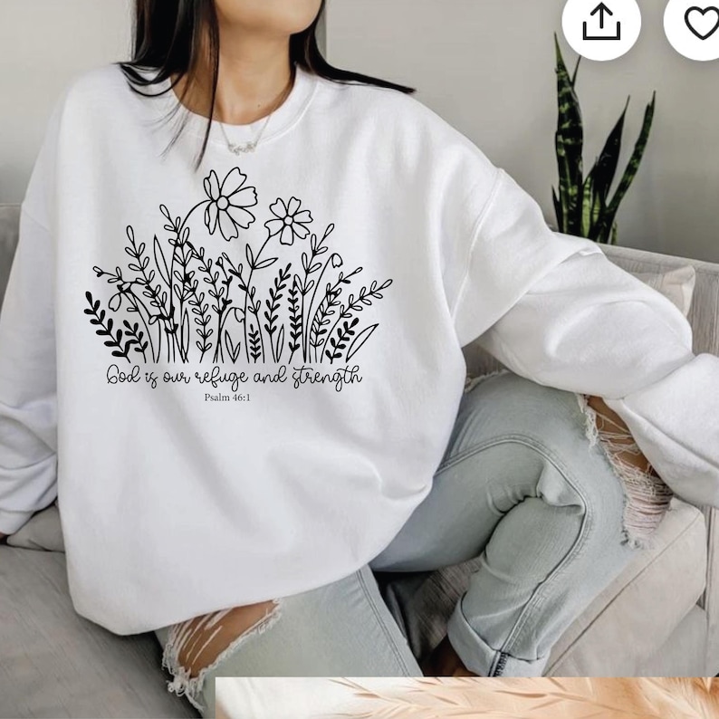 May include: White sweatshirt featuring a black floral design with the text "God is our refuge and strength" and "Psalm 46:1". The sweatshirt has a crew neck and long sleeves. The design includes flowers, leaves, and stems.