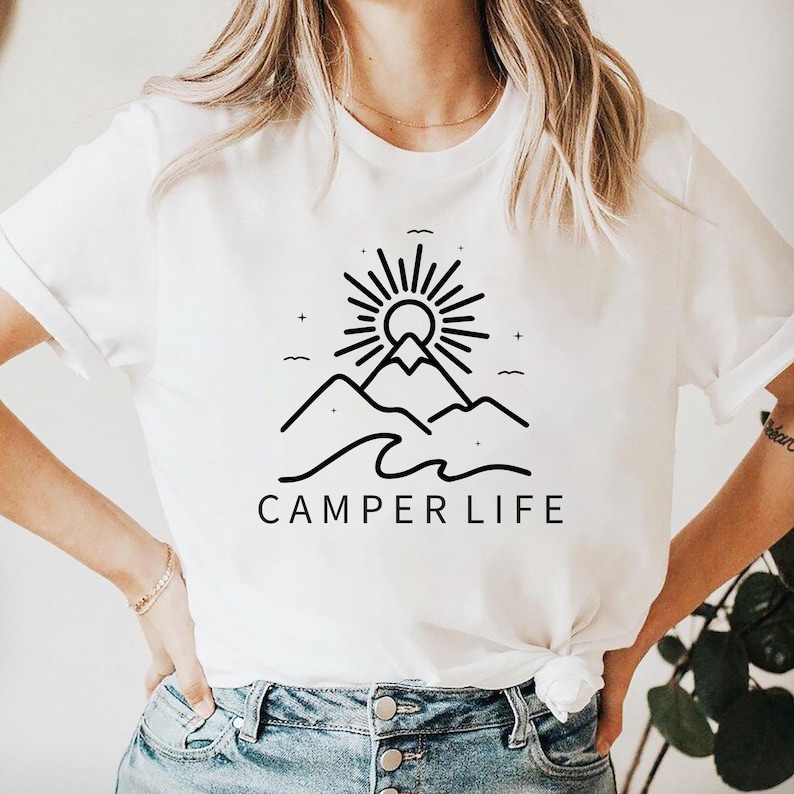 May include: White t-shirt with a black graphic of a mountain range, sun, and waves. The text "CAMPER LIFE" is below the graphic. The shirt is made of a soft material.