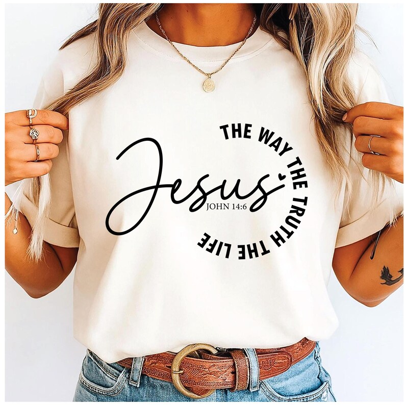 Puede incluir: Camiseta color crema con la palabra "Jesus" en escritura negra fluida. El texto "THE WAY THE TRUTH THE LIFE" rodea la palabra "Jesus". La camiseta tambi&eacute;n incluye el texto "JOHN 14:6".