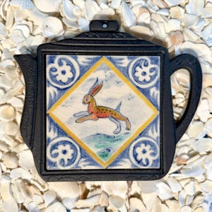 May include: Black teapot-shaped trivet with a ceramic tile insert. The tile features a running rabbit in orange and yellow, set against a blue and white floral pattern. The trivet is designed to protect surfaces from hot items.