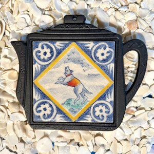 May include: Black cast iron teapot-shaped trivet with a decorative tile insert. The tile features a dog wearing a vest, set within a blue and yellow geometric design. The trivet has a handle and a hole for hanging.