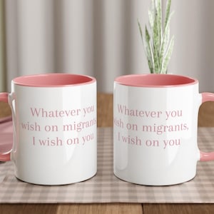 Könnte beinhalten: Zwei weiße Keramikbecher mit rosa Innenraum und Griffen. Die Becher tragen den Text "Whatever you wish on migrants, I wish on you" in hellrosa. Die Becher stehen auf einer karierten Oberfläche.