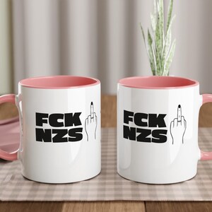 May include: Two white ceramic mugs with pink interiors and handles. Each mug features the text "FCK NZS" in bold black letters, accompanied by a hand giving the middle finger. The mugs are on a checkered tablecloth.