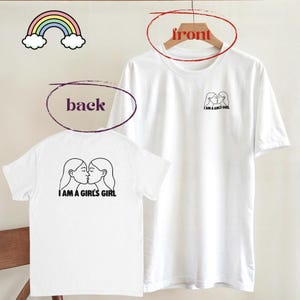 Peut inclure: T-shirts blancs avec un design minimaliste. Le devant du t-shirt présente un petit dessin au trait de deux figures s'embrassant, avec le texte "I AM A GIRL'S GIRL" au dos. Un arc-en-ciel est dans le coin supérieur gauche.