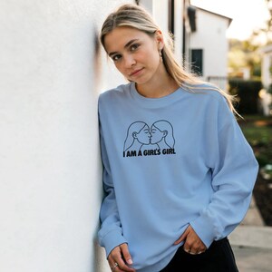 Könnte beinhalten: Hellblaues Sweatshirt mit langen Ärmeln. Auf der Vorderseite befindet sich eine schwarze Strichzeichnung von zwei küssenden Frauen mit dem Text "I AM A GIRL'S GIRL" darunter. Das Sweatshirt ist aus weichem Material.