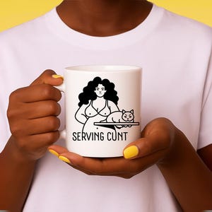 Könnte beinhalten: Weiße Keramik-Tasse mit einer schwarzen Grafik einer Frau und einer Katze, mit dem Text "SERVING CUNT". Die Tasse wird von einer Person gehalten. Die Tasse hat eine Standardgröße für Kaffee oder Tee.
