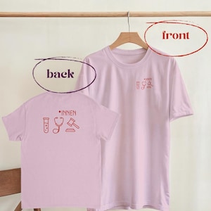 Könnte beinhalten: Zwei hellrosa T-Shirts hängen an einem Holzkleiderbügel. Die Vorderseite des Shirts auf der rechten Seite hat eine kleine rote Grafik eines Bechers, eines Stethoskops und eines Hammers. Auf der Rückseite des Shirts auf der linken Seite steht das Wort "INNEN" über denselben Grafiken.