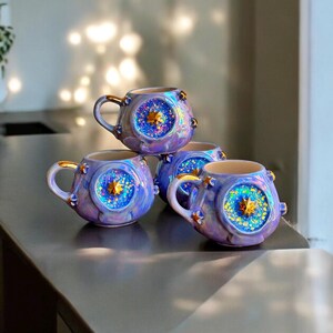May include: Four ceramic mugs with a celestial design. The mugs are a gradient of purple and blue with gold accents on the handles and rims. Each mug features a central starburst design with glitter.