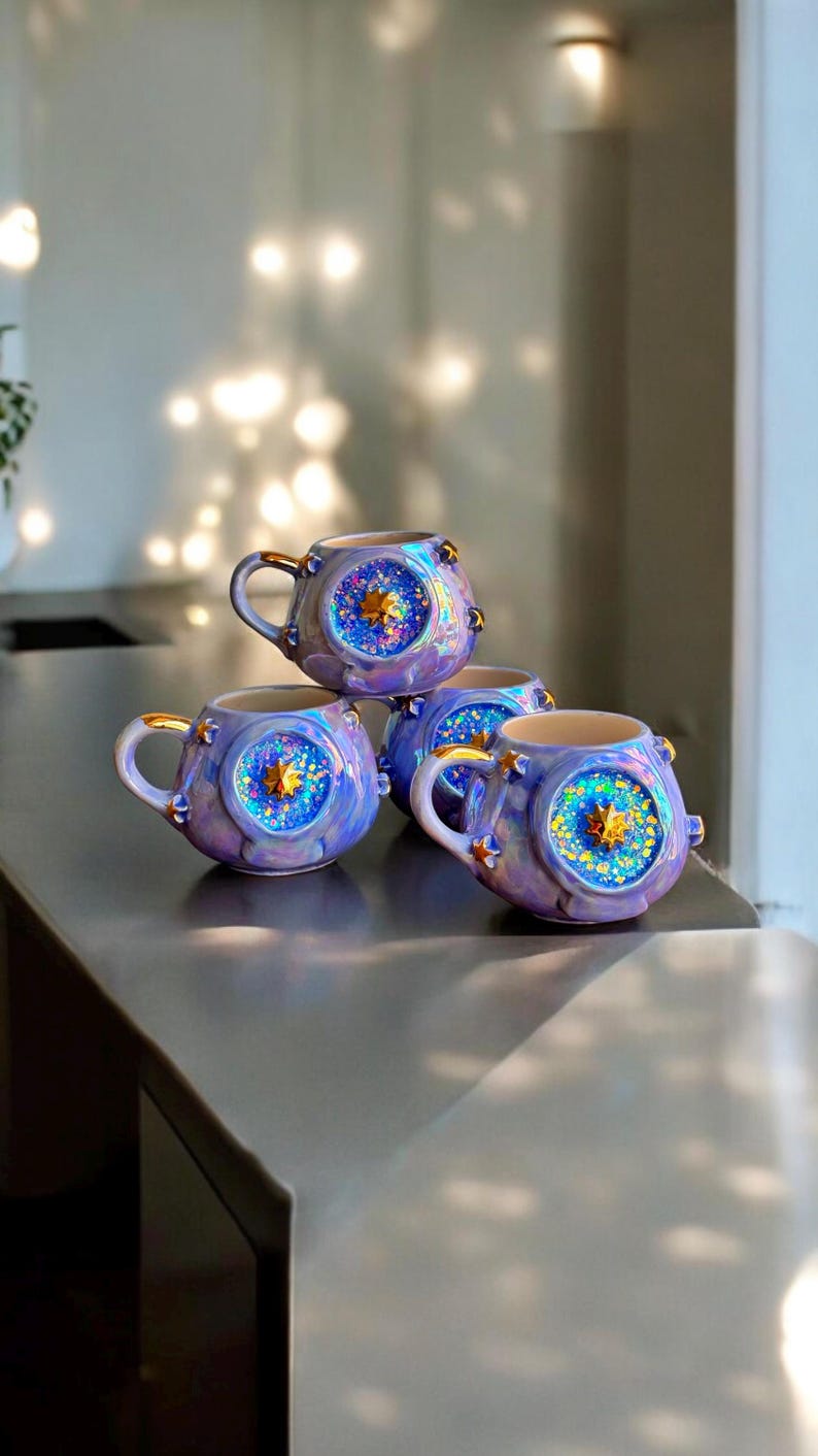 May include: Four ceramic mugs with a celestial design. The mugs are a gradient of purple and blue with gold accents on the handles and rims. Each mug features a central starburst design with glitter.