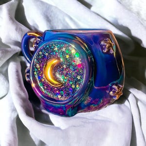 May include: A blue iridescent ceramic mug with a gold handle and star accents. The mug features a circular design with a gold crescent moon and colorful glitter. The mug is on a white surface.