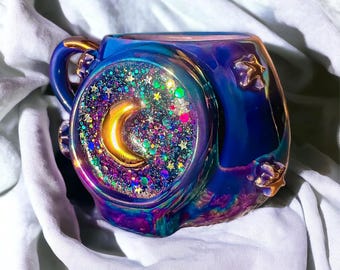 May include: A blue iridescent ceramic mug with a gold handle and star accents. The mug features a circular design with a gold crescent moon and colorful glitter. The mug is on a white surface.