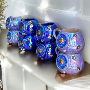 May include: A collection of iridescent blue ceramic mugs with star and moon designs. Each mug has a gold handle and a decorative front with a celestial theme. The mugs are stacked on a shelf.