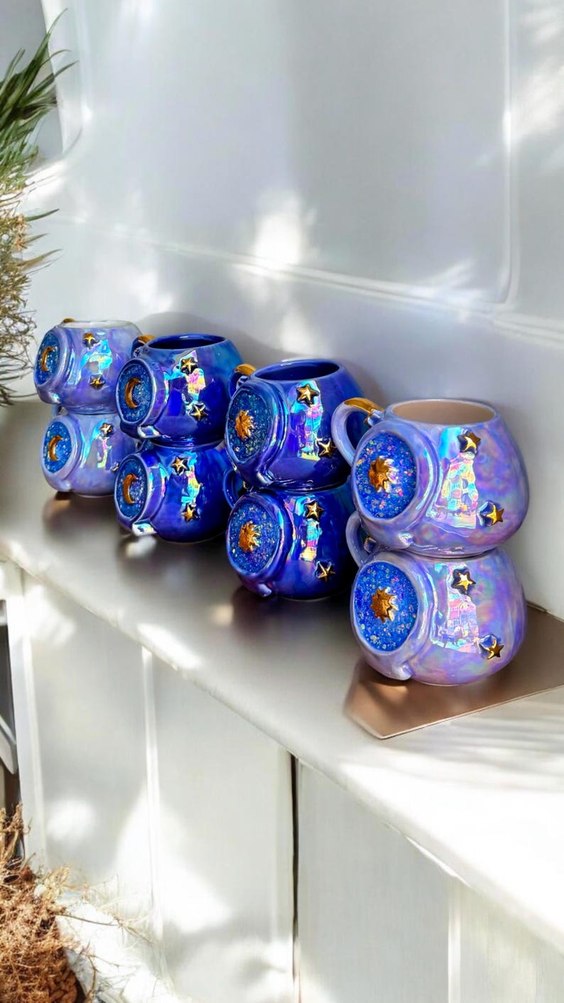 May include: A collection of iridescent blue ceramic mugs with star and moon designs. Each mug has a gold handle and a decorative front with a celestial theme. The mugs are stacked on a shelf.