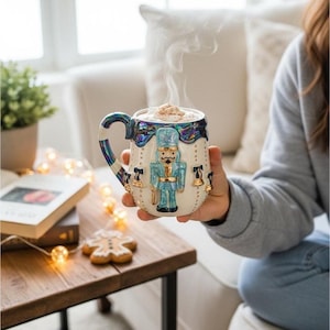 May include: A decorative ceramic mug with a blue and gold handle and rim, featuring a blue nutcracker soldier design. The mug is filled with a hot drink, and steam rises from the top. Gold bells and a blue bow decorate the mug.