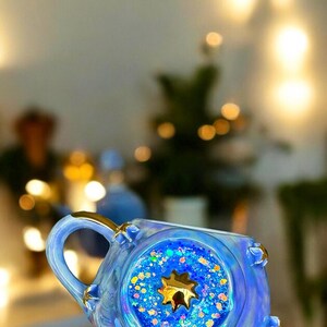 May include: A blue ceramic mug with a gold handle and accents. The mug features a circular design filled with blue glitter and a gold star. The mug is iridescent and has a unique, decorative design.