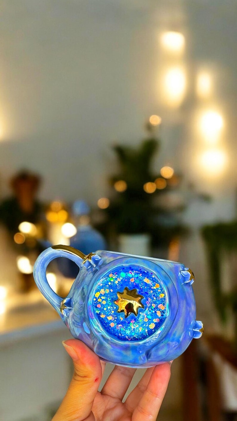 May include: A blue ceramic mug with a gold handle and accents. The mug features a circular design filled with blue glitter and a gold star. The mug is iridescent and has a unique, decorative design.