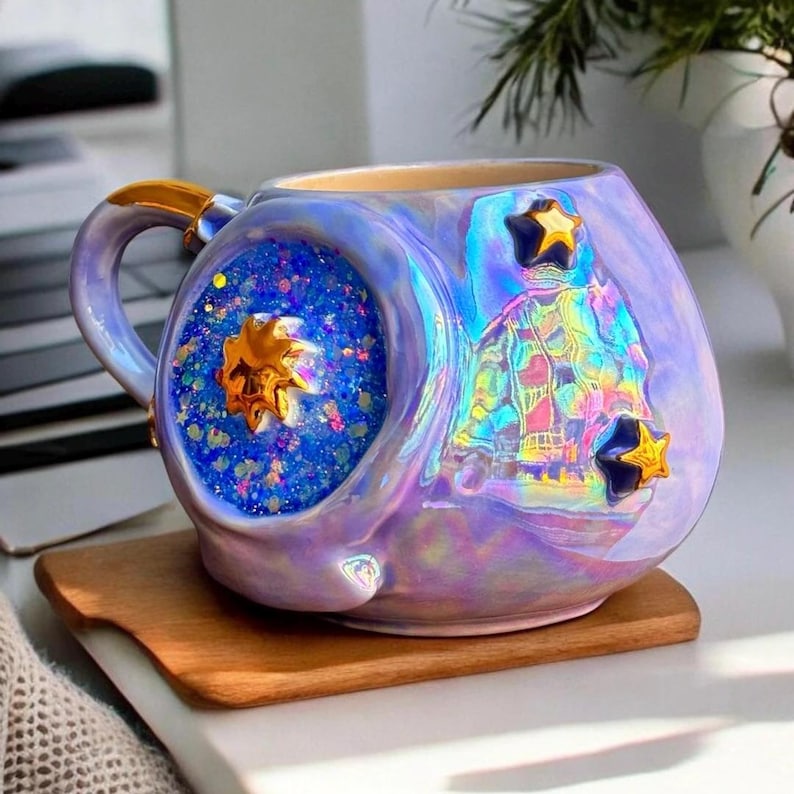 May include: Iridescent ceramic mug with a unique design. The mug features a blue glitter-filled sphere with a gold star, and a colorful, reflective design on the side. The handle has a gold accent. The mug sits on a wooden coaster.