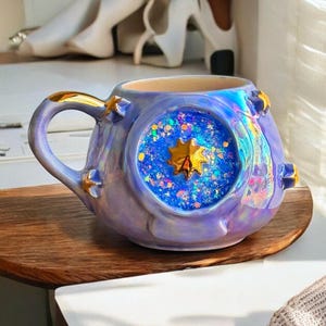 May include: A ceramic mug with a celestial theme. The mug is a lavender color with gold accents and a large, round window filled with blue glitter and a gold star. The handle and star details are gold. The mug sits on a wooden surface.
